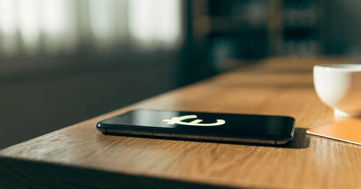 Zwei Euro-Scheine und ein Smartphone liegen auf einem Holztisch neben einer Tasse, im Hintergrund ein unscharfes Fenster.