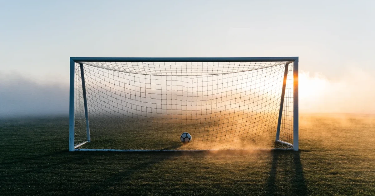 Ein Fußballtor mit gespanntem Netz steht auf einem leeren Rasenplatz, davor ein einzelner Ball, Morgendunst liegt über dem Feld.