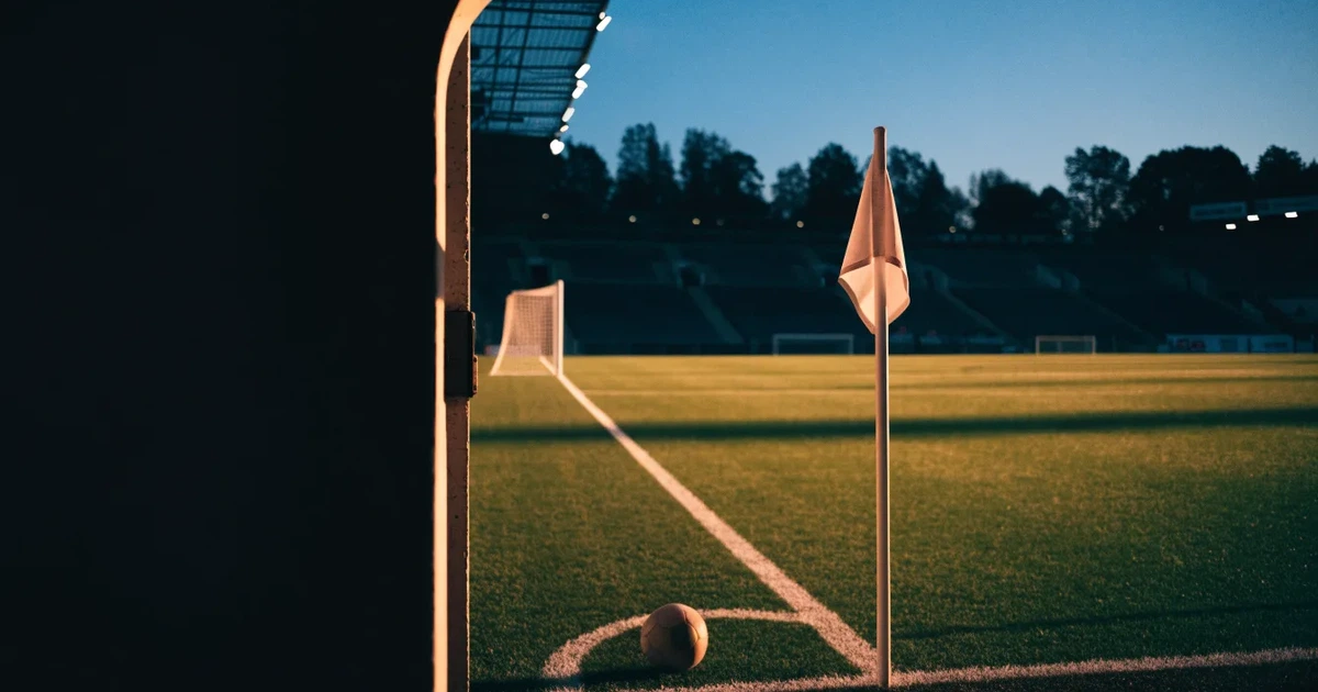 Blick durch ein Stadiontor auf einen grünen Rasen im Abendlicht, ein Ball liegt an der Eckfahne, die Ränge sind leer.