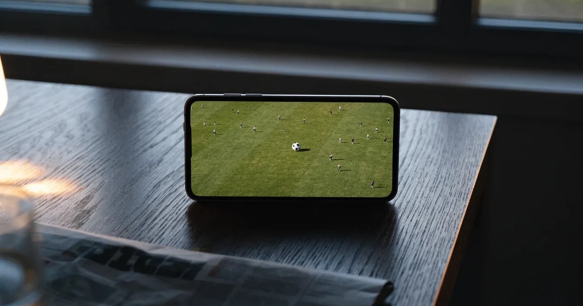 Ein Fußballstadion unter Flutlicht aus großer Entfernung, davor ein dunkler Parkplatz und ein heller Abendhimmel.