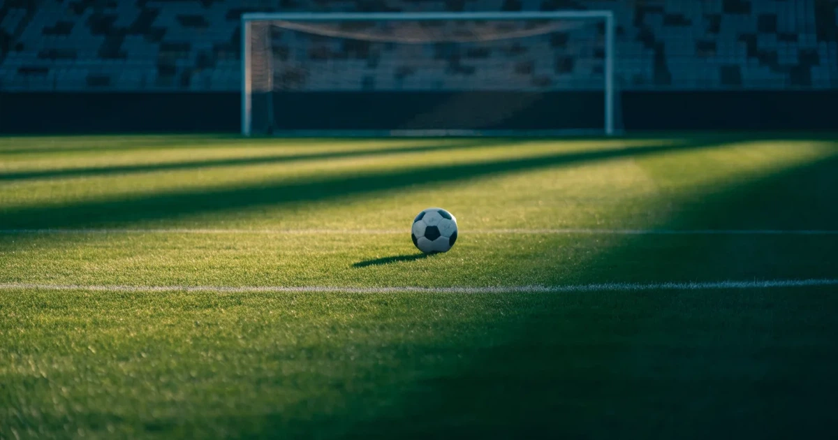 Ein Fußballplatz mit frisch gemähtem Rasen im Abendlicht, ein Ball liegt im Strafraum, im Hintergrund unscharf eine leere Tribüne.