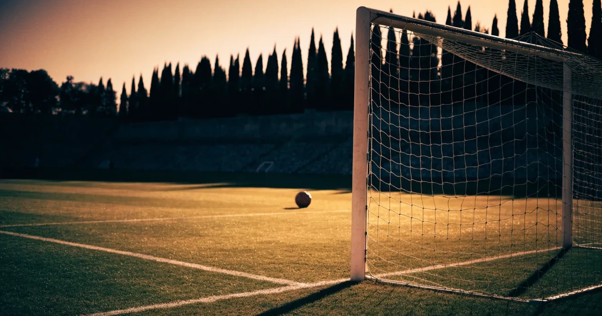 Ein Fußballtor mit Netz auf einem leeren Platz im Abendlicht, der Rasen frisch gemäht, im Hintergrund Zypressen.