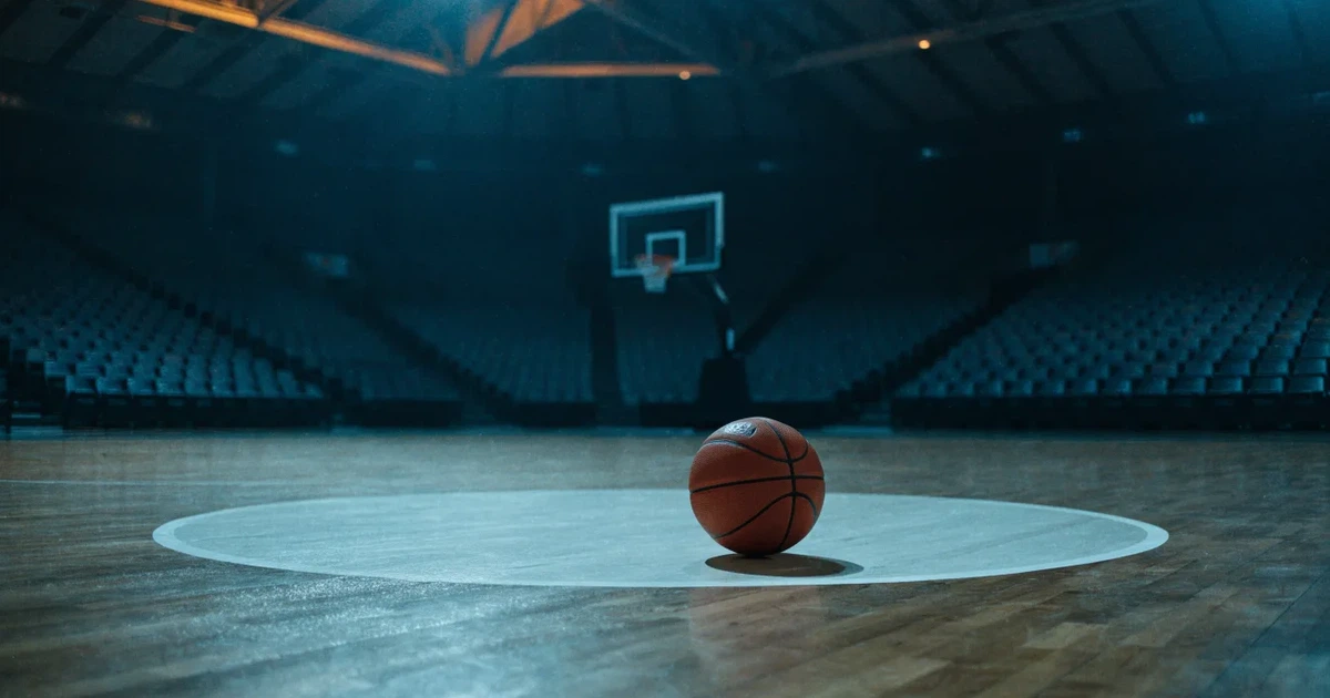 Ein Basketball liegt auf dem Parkettboden einer leeren Arena, Scheinwerfer werfen Licht auf die Mittellinie.