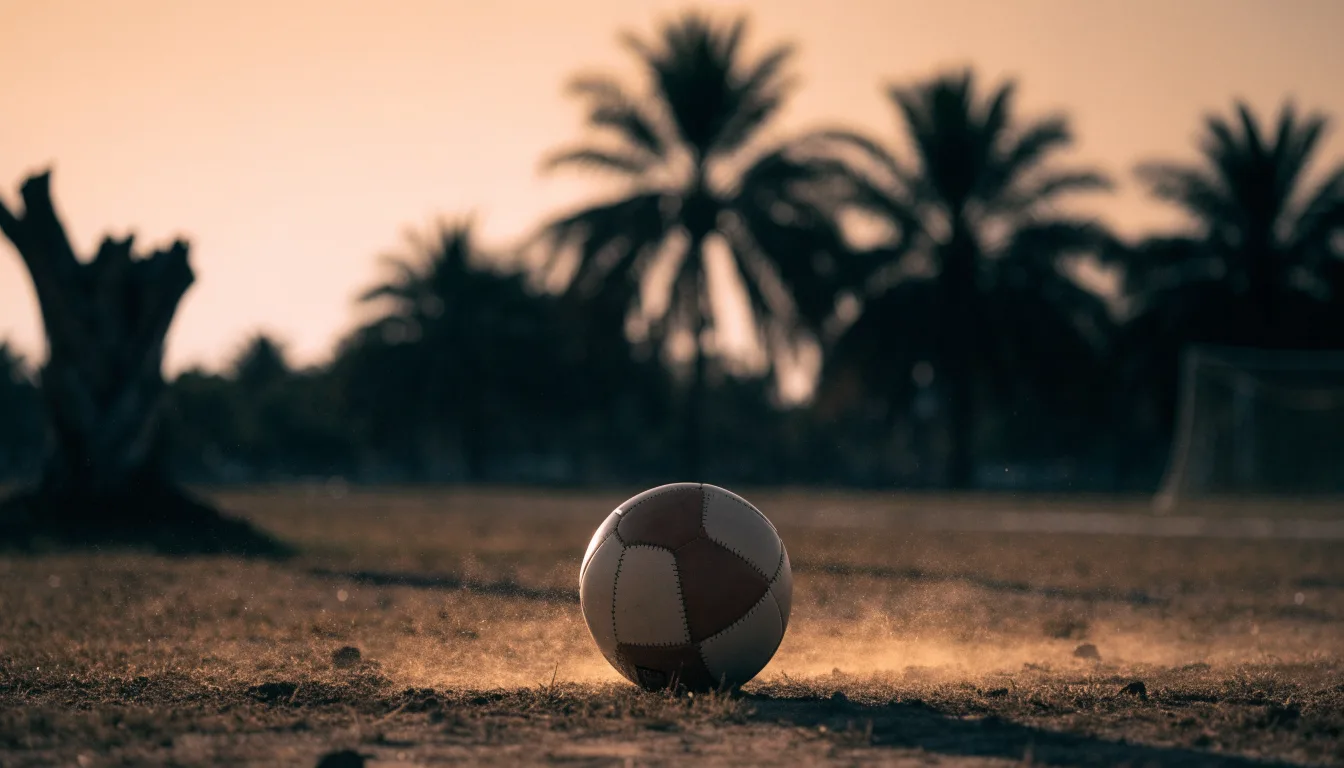 Ein Fußball liegt auf einem staubigen Bolzplatz mit Palmen im Hintergrund in warmem Abendlicht.