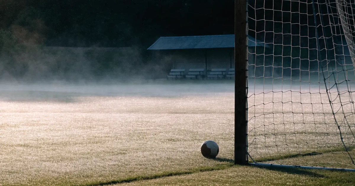 Ein Fußballtor mit Netz steht im Morgennebel auf einem leeren Rasenplatz, im Hintergrund die Silhouette einer kleinen Tribüne.