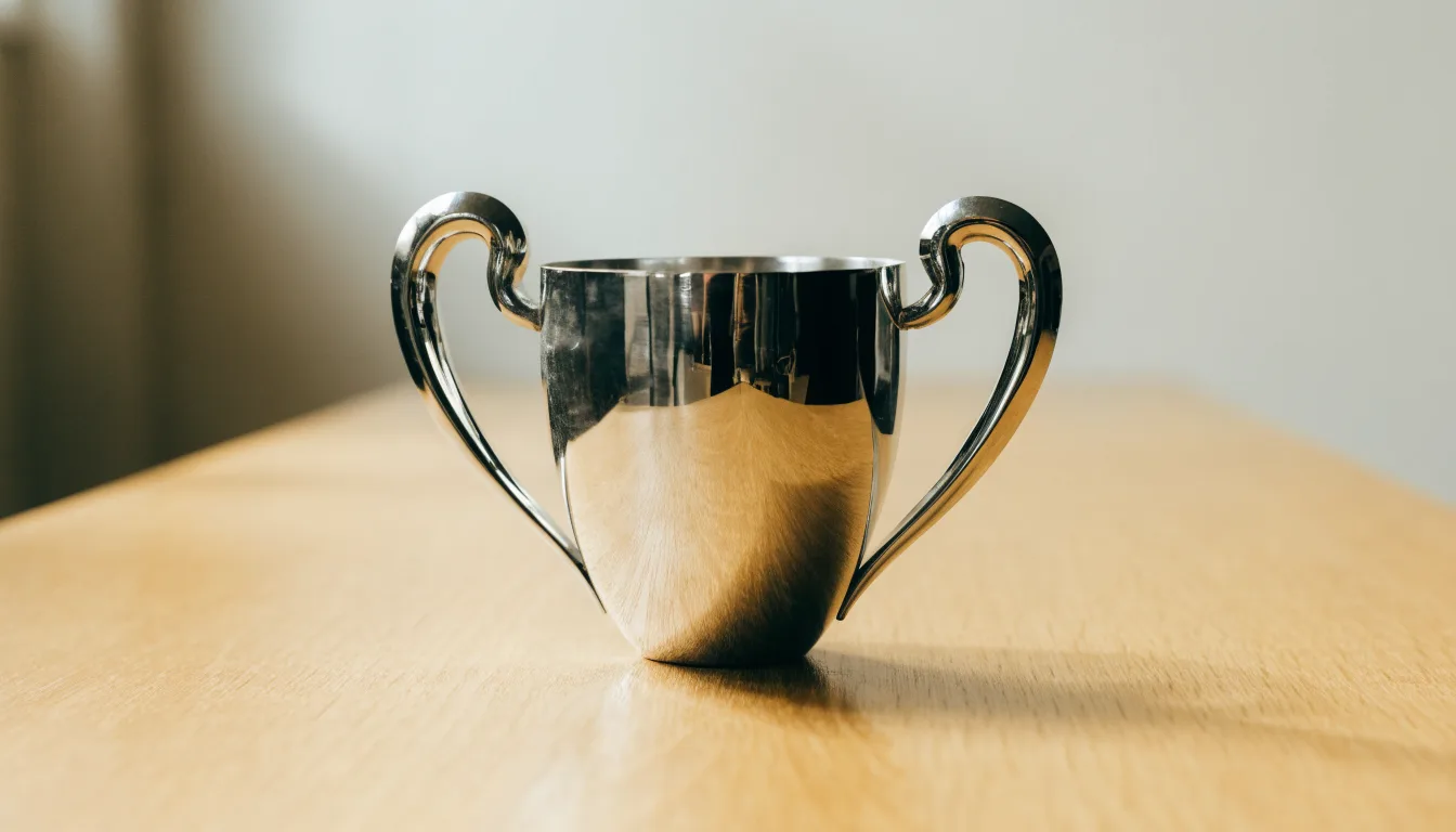 Ein Pokal ohne Gravur steht allein auf einem Holztisch vor einer hellen Wand, davon fällt ein weicher Schatten.