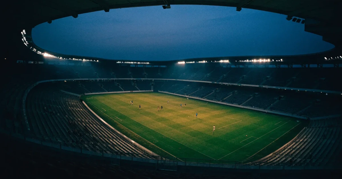 Ein Fußballstadion am Abend unter Flutlicht aus der Ferne gesehen, die Tribünen sind leicht beleuchtet und der Himmel dunkelblau.