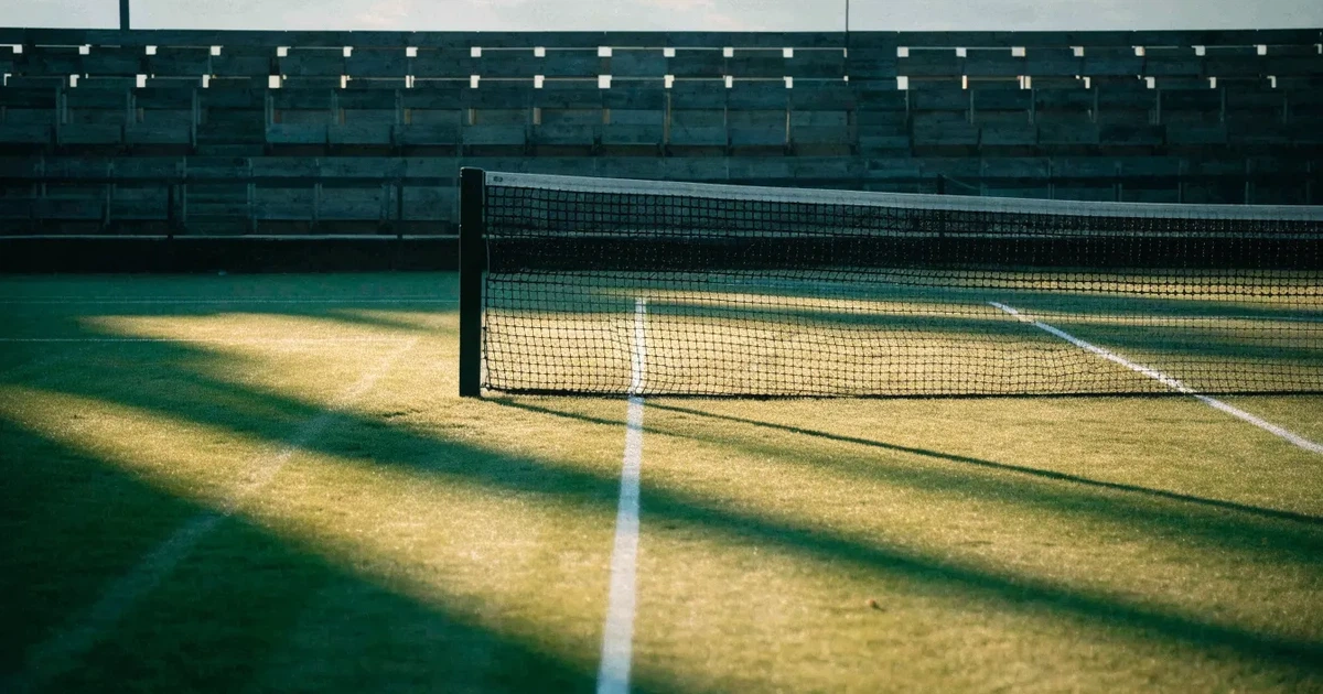 Ein Tennisplatz mit Rasenbelag im Morgenlicht, Netz und weiße Linien, im Hintergrund eine ruhige Tribüne.